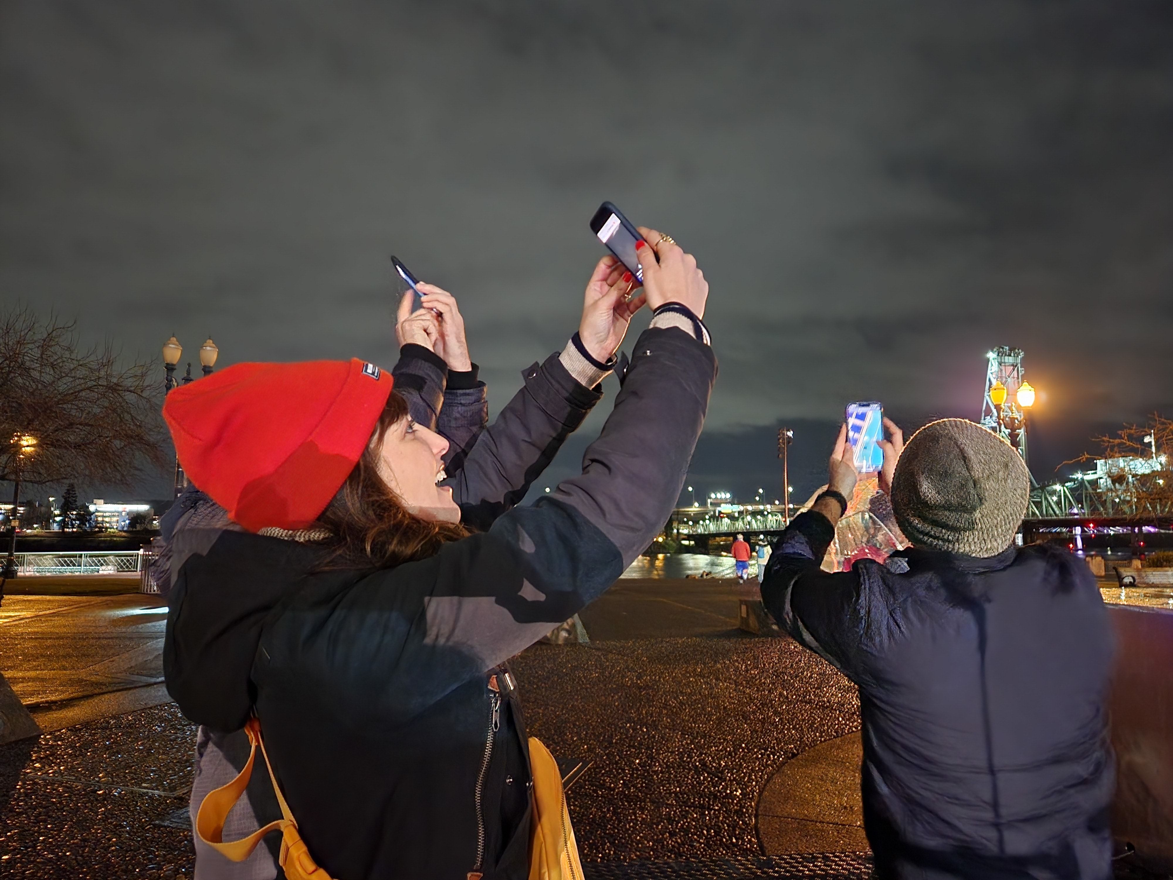 Three people holding up their mobile devices towards the sky, outdoors at night in Portland, Oregon.